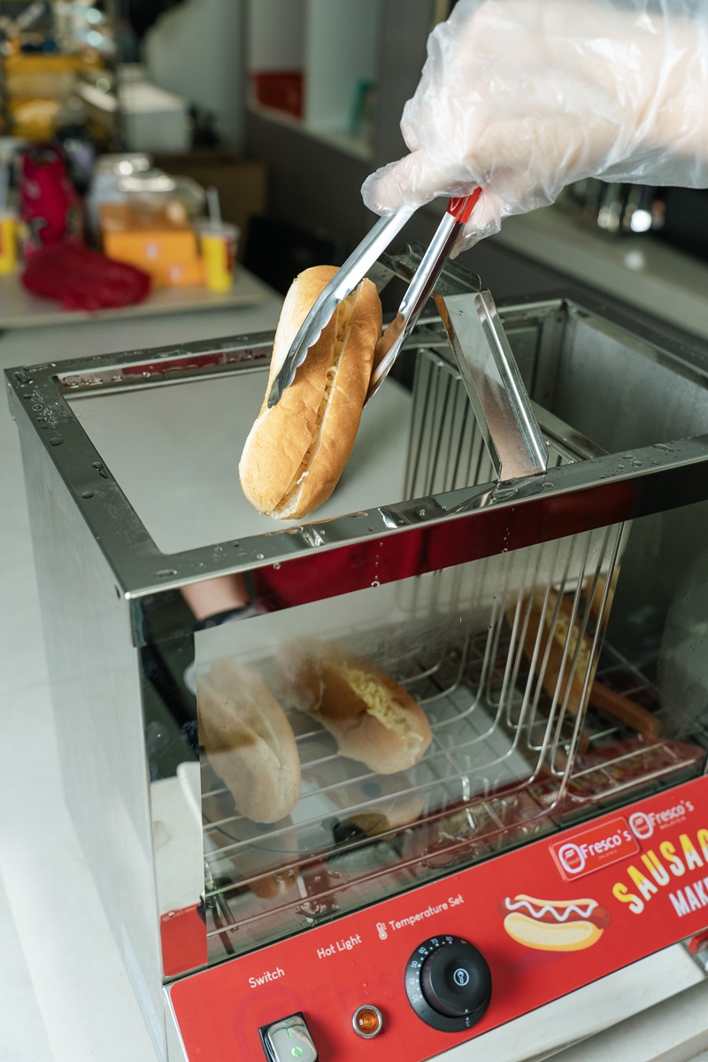 A gloved hand uses tongs to remove a steamed hotdog bun from a commercial sausage warmer with visible temperature controls, perfect for any Hotdog steamer Malaysia setup.
