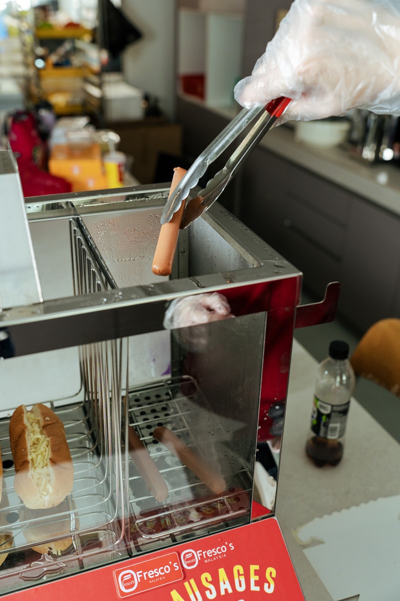 A gloved hand uses tongs to place a steamed hotdog into a hot dog machine next to a prepared hot dog bun in a kitchen setting.