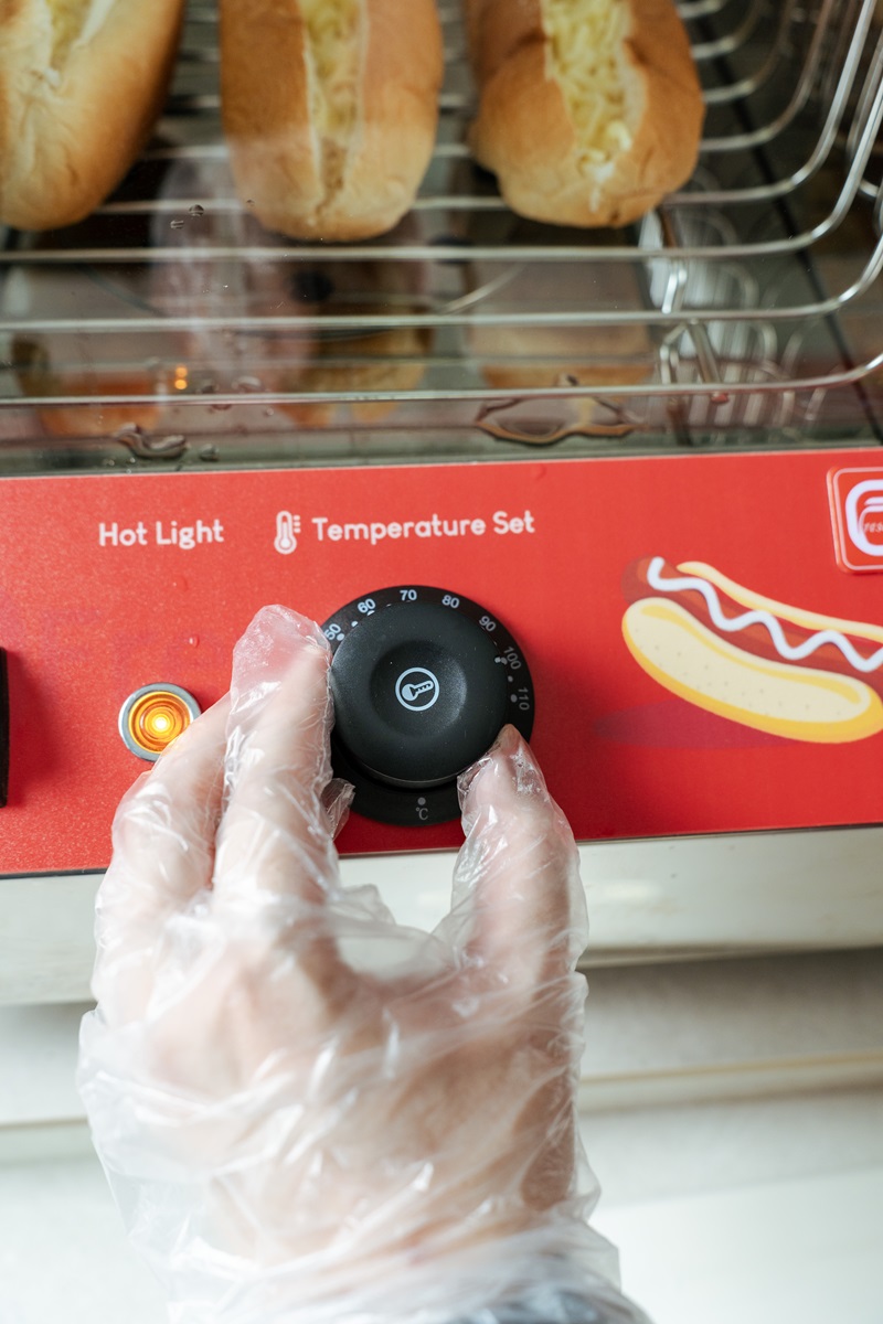 A gloved hand adjusts the temperature dial on a hot dog warmer with several steamed hotdog Malaysia-style treats inside.