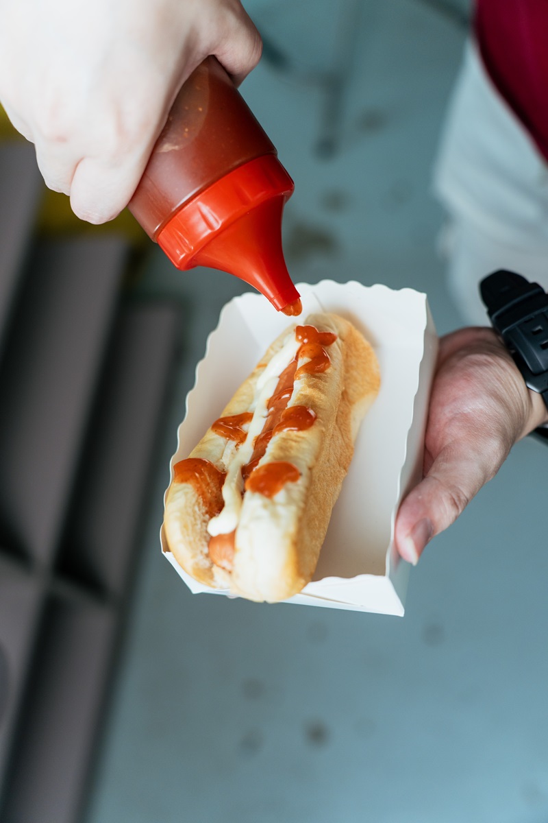 A person holds a hot dog in a paper tray while squeezing ketchup from a bottle onto it, showcasing an essential step in how to make hotdogs at home.