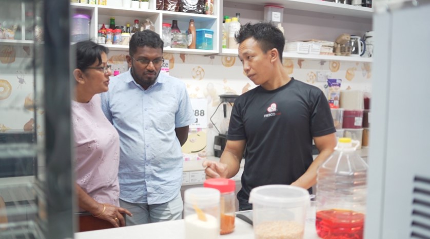 Three people are standing in a kitchen, talking. One man is explaining something while gesturing toward a Fresco popcorn maker on the counter, surrounded by various kitchen items and containers on the shelves around them.