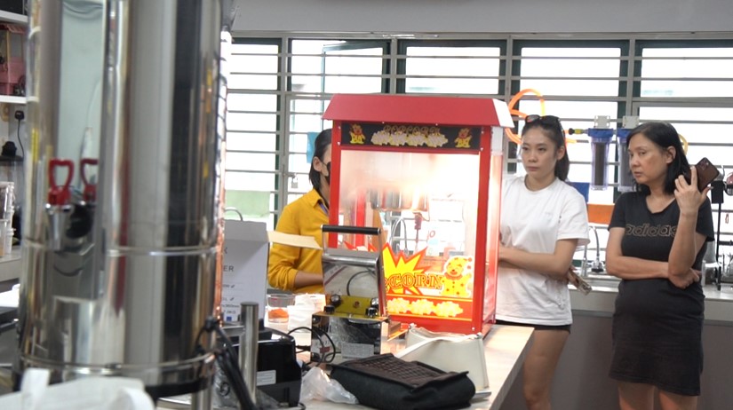 Two women stand next to a Popcorn machine Malaysia in a modern room with large windows and various equipment on the counters, discussing how to make cinema popcorn.