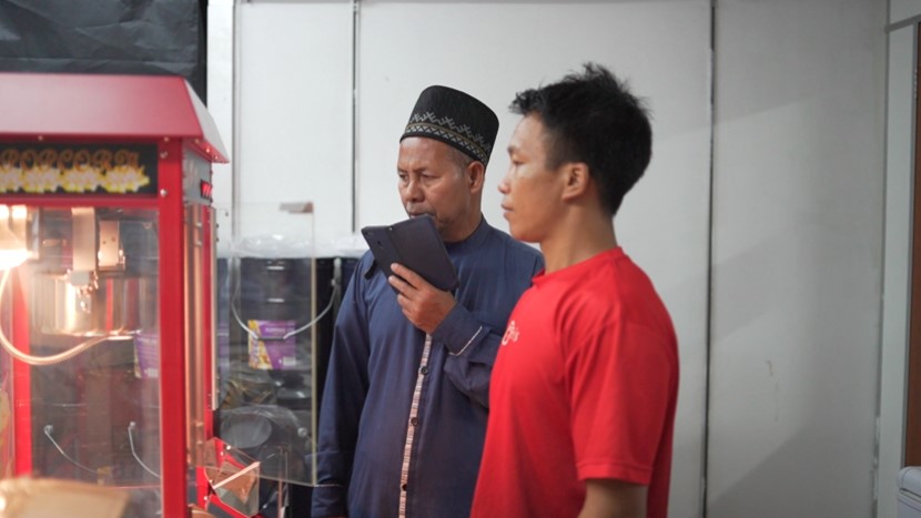 Two men stand indoors; one in a blue traditional outfit holding a tablet, the other in a red T-shirt, both looking at a popcorn machine Malaysia, possibly discussing how to make cinema popcorn.