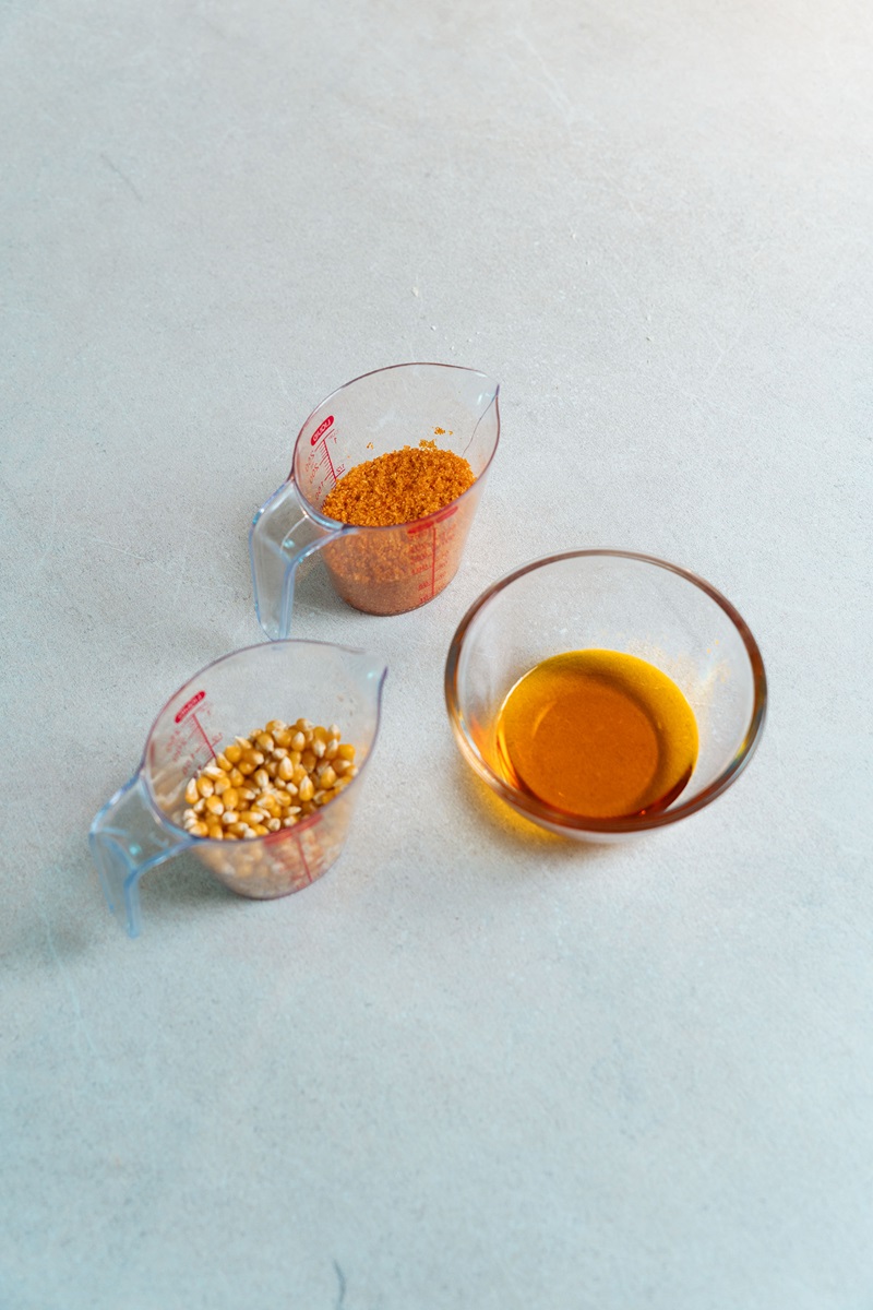 Two measuring cups with popcorn kernels and cheese powder, and a glass bowl with melted butter on a light surface—perfect ingredients for learning how to make cinema popcorn at home or for your commercial popcorn machine.