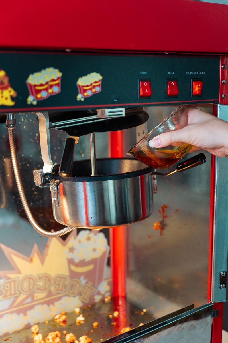 A hand pours oil from a glass container into the metal kettle of a popcorn machine, showing how to make cinema popcorn just like in theaters.