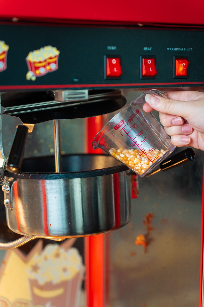 A hand pours popcorn kernels from a measuring cup into the kettle of a red Fresco popcorn maker, showing how to make cinema popcorn at home.