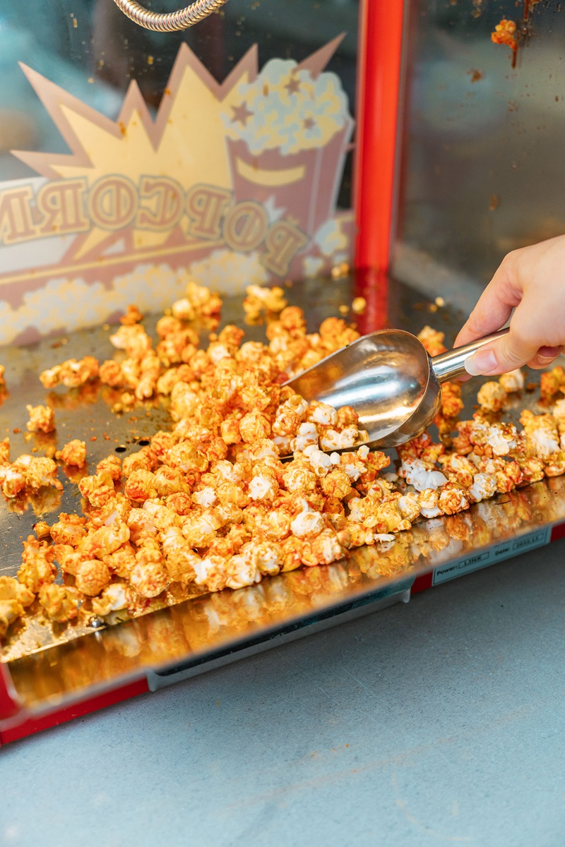 A hand uses a metal scoop to serve caramel popcorn from a retro Fresco popcorn maker, adding a nostalgic touch to any snack time.