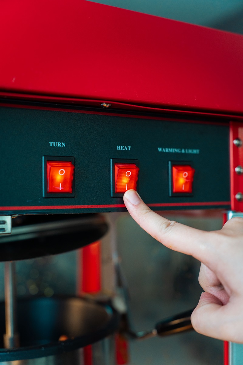 A hand presses the "Heat" switch on a red popcorn machine control panel, showing how to make cinema popcorn just like in theaters. This Popcorn machine Malaysia model also features "Turn" and "Warming Light" switches for perfect results.