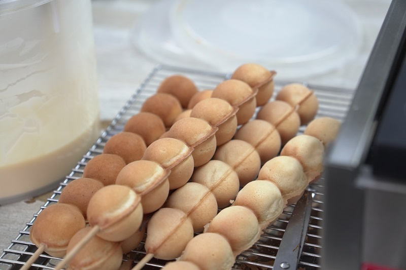 Several skewers of round, golden brown waffle balls rest on a metal cooling rack beside a container of batter, perfect for anyone looking for an easy waffle balls recipe.