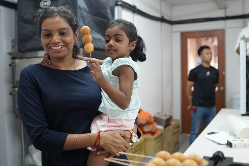 A woman holds a young girl who is enjoying crispy waffle balls on a skewer in a room with another person and some boxes in the background.