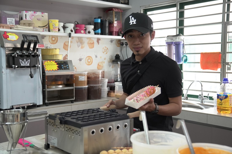 A man in a black shirt and LA cap holds a tray of snacks in a kitchen equipped with appliances, containers, baking supplies, and a waffle ball machine for trying out his favorite waffle balls recipe.