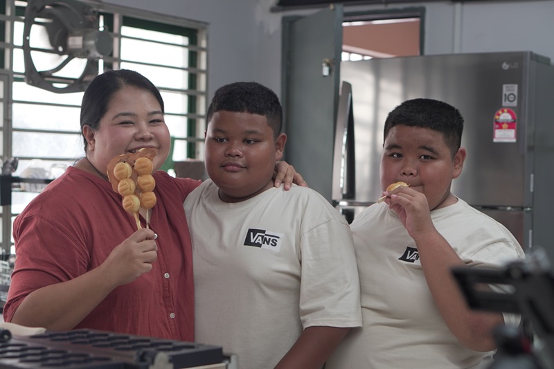 A woman stands beside two boys in a kitchen. The woman holds skewers with crispy waffle balls, while one boy also enjoys a skewer. All three are facing the camera, showcasing their delicious waffle ball treats.