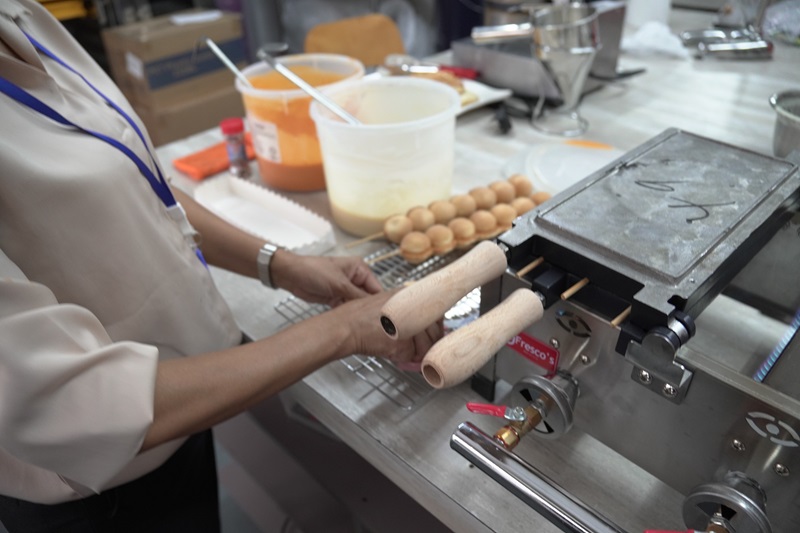 A person operates a waffle ball machine at a kitchen workstation with bowls of batter, a cooling rack, and finished crispy waffle balls nearby.