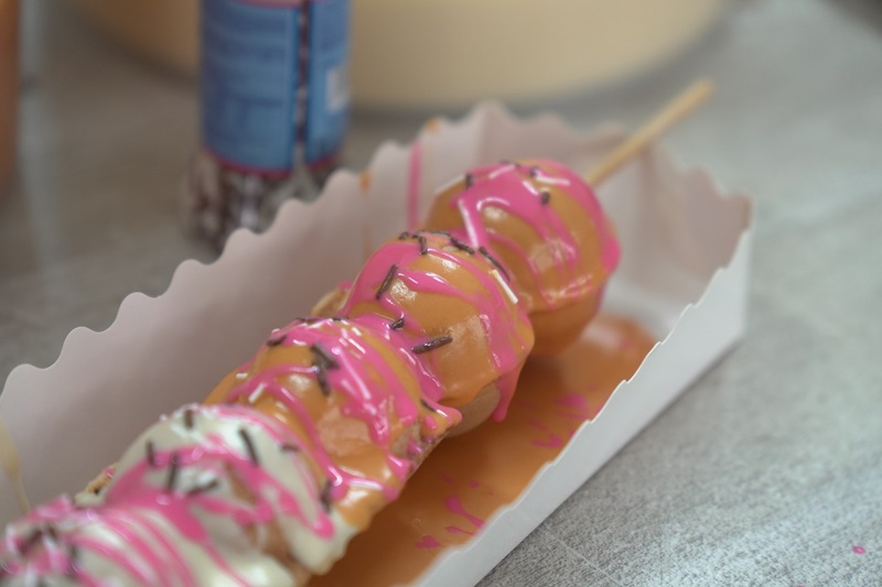 A skewer of waffles, made with a waffle ball machine and drizzled with pink and white icing and chocolate sprinkles, served in a white paper tray.