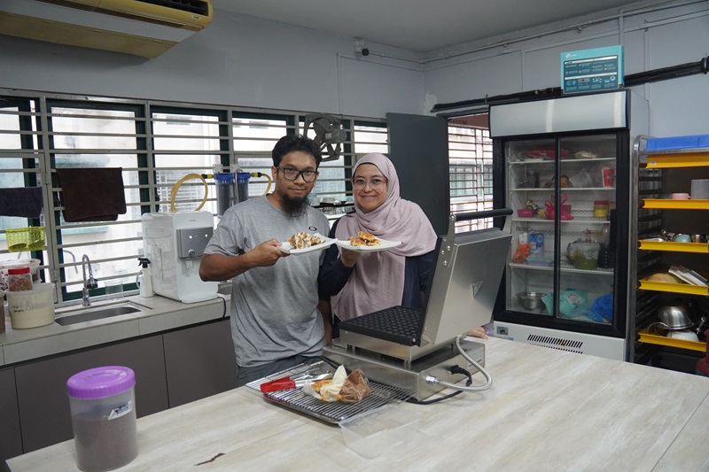 Two people standing in a kitchen hold plates of food and smile at the camera, with a Professional Croffle Maker and shelves filled with kitchen appliances visible in the background.