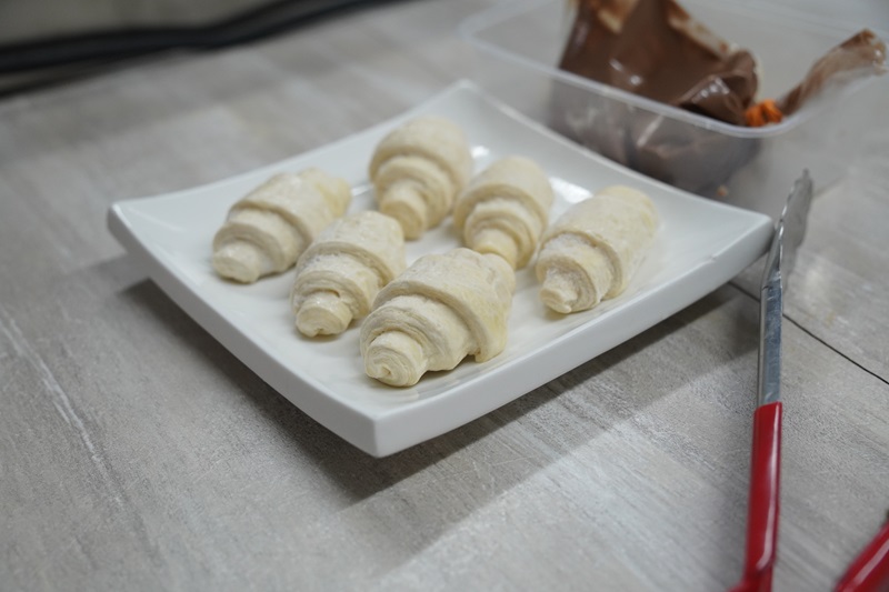 Six uncooked croissant rolls are arranged on a square white plate, ready to be transformed into Croissant Waffles using your Professional Croffle Maker, with a container of chocolate spread and red-handled tongs nearby.