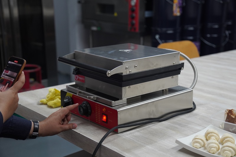 A person adjusts the settings on a stainless steel Croffle Maker on a table, with pastries, including a Croissant Waffle, and a smartphone visible nearby.