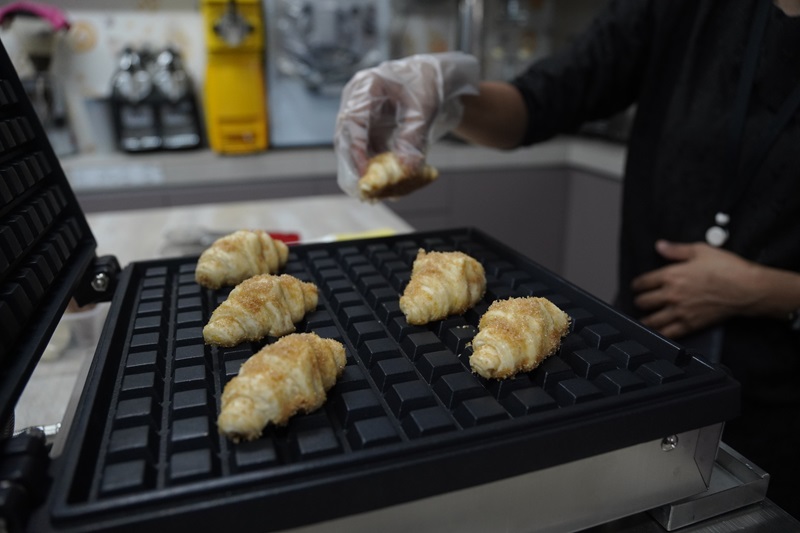 A person wearing a glove places croissants onto a Croffle Maker in a kitchen setting, preparing delicious croissant waffles.