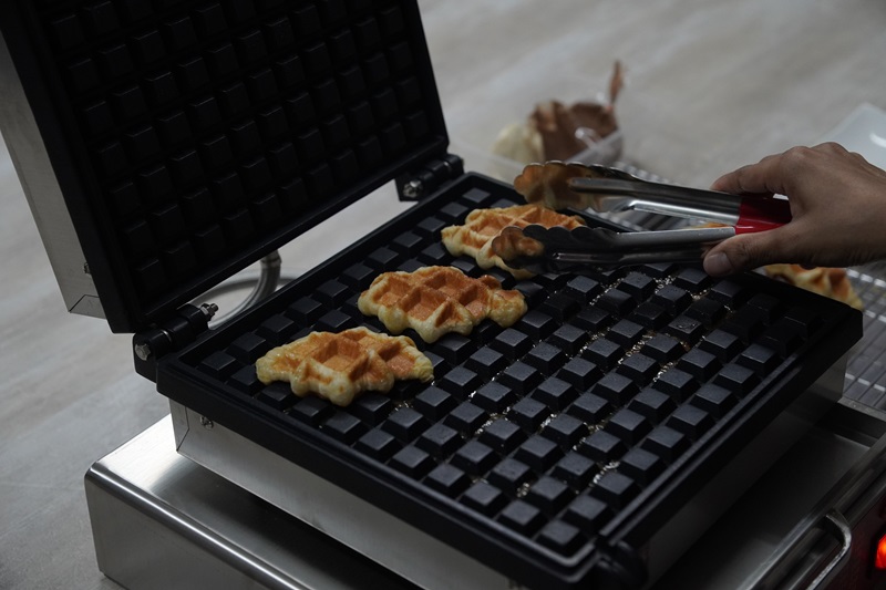A person uses tongs to remove freshly cooked Croissant Waffles from a professional croffle maker.