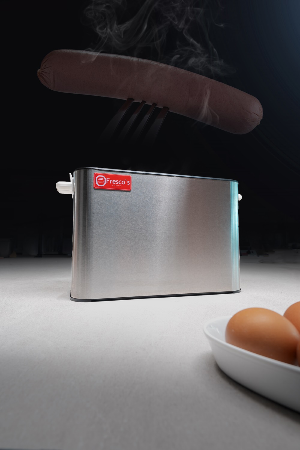 A hot dog is being lifted by a fork above a stainless steel toaster on a counter, with two eggs on a dish in the foreground—perfect inspiration for planning your next breakfast or searching for the ideal waffle balls recipe.