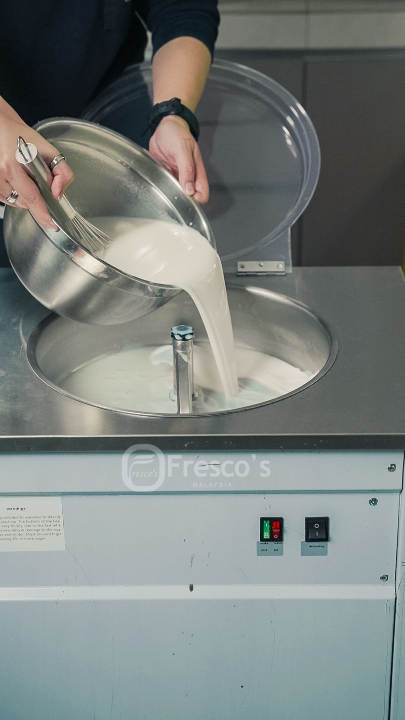 A person pours a large bowl of white liquid into an industrial ice cream and sorbet maker labeled "Fresco's Malaysia," showing how to make frozen dessert.