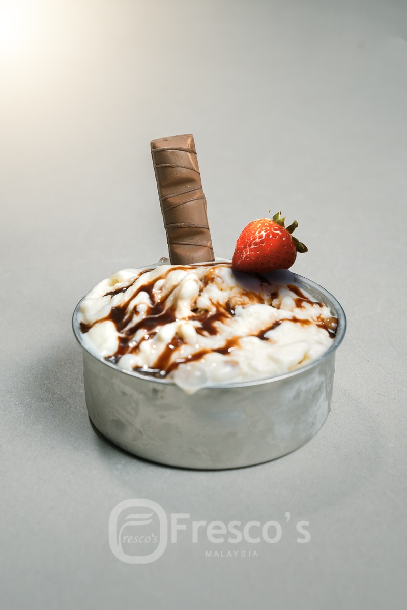 A metal bowl of vanilla ice cream, topped with chocolate syrup, a chocolate wafer stick, and a fresh strawberry sits on a gray surface—perfect for serving desserts made in your favorite ice cream and sorbet maker.