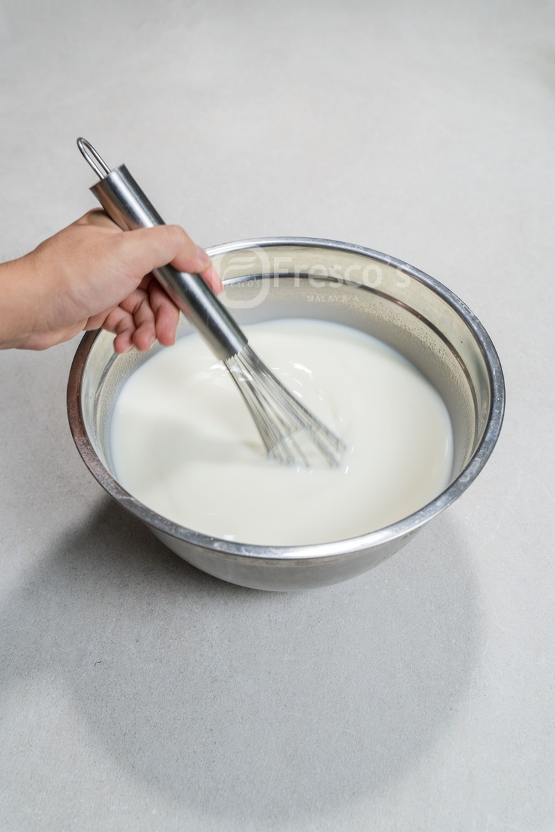 A hand whisking a white liquid, likely milk or batter, in a stainless steel bowl on a light-colored surface—perfect for preparing mixtures before using a sorbet machine or learning how to make sorbet at home.