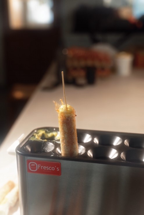 A cooked corndog on a stick stands upright in a metal corndog maker labeled "Fresco's" on a kitchen counter, next to a waffle ball machine perfect for making crispy waffle balls.