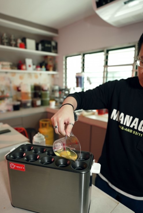 A person pours a yellow liquid from a measuring cup into a muffin tin on a kitchen counter, prepping the batter for crispy waffle balls.