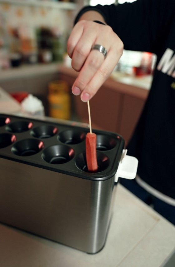 A person places a hot dog on a stick into a compartment of a metallic kitchen appliance, likely a waffle ball machine, preparing to make a creative snack.