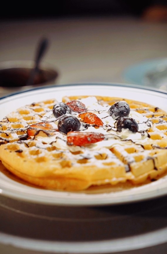 A plate with a waffle topped with whipped cream, sliced strawberries, blueberries, powdered sugar, and chocolate syrup—perfect inspiration if you're searching for the Best Waffle Recipe or curious about how to make waffle at home.