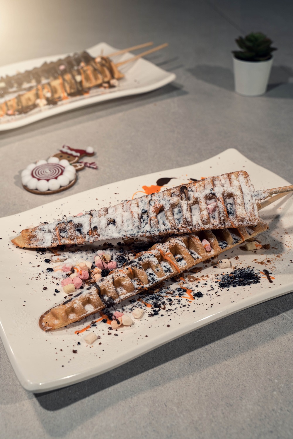 Two waffle sticks made with a waffle stick maker rest on a rectangular white plate, dusted with powdered sugar and cocoa, and garnished with colorful candy bits. A potted plant and another plate are visible in the background.