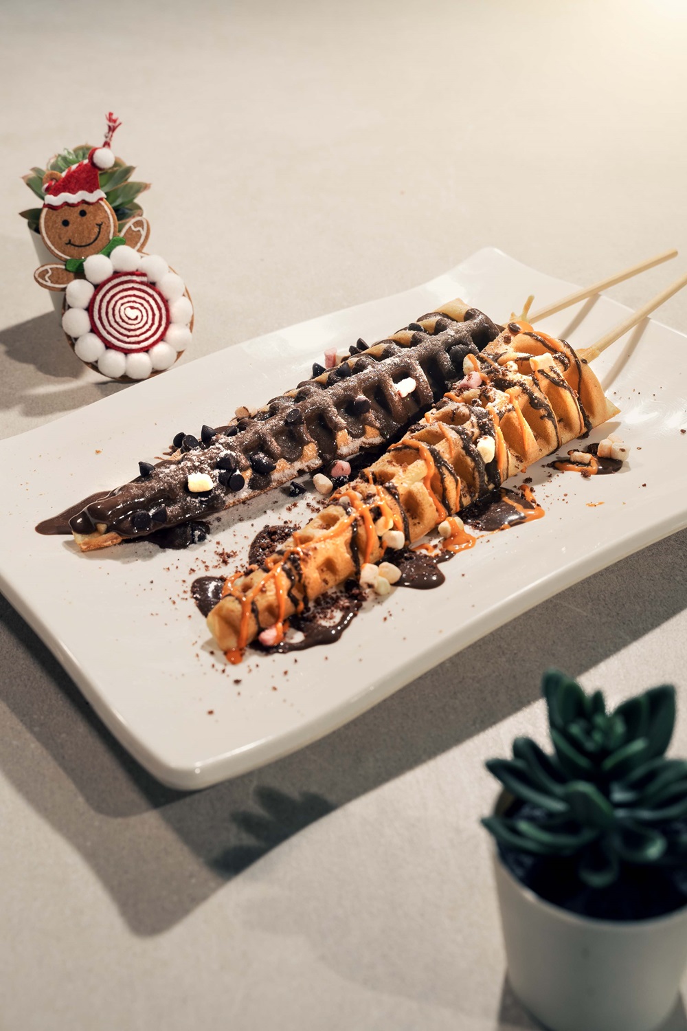 Two skewered waffle sticks, crafted with a waffle machine Malaysia, are drizzled with chocolate and toppings on a white plate; a decorative Christmas figure and small potted plant add a festive touch nearby.