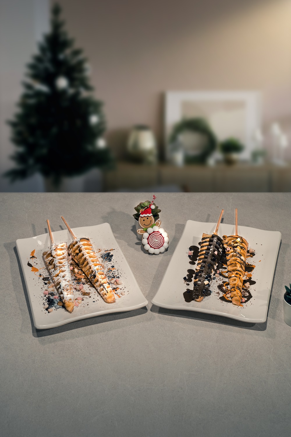 Two rectangular plates hold churros on sticks, topped with various sauces and sprinkles, on a table with a small holiday decoration; a Christmas tree is blurred in the background, evoking festive fun with treats made using a waffle stick maker.