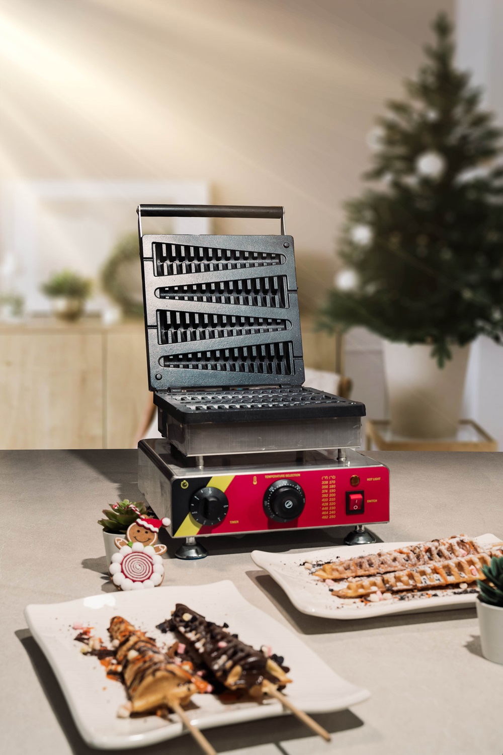 A commercial waffle stick maker sits on a countertop with two plates of decorated Christmas Tree Waffle sticks in front; festive decor and a blurred Christmas tree are in the background.