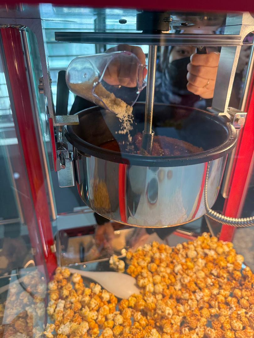 During a popcorn machine demo, a person pours popcorn seasoning into the machine, with freshly popped caramel popcorn visible in the tray below.