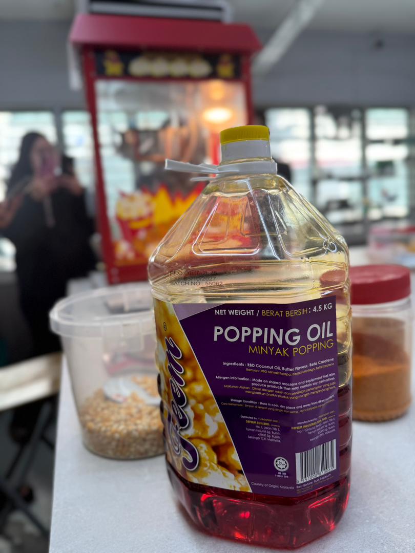 A large plastic bottle of popping oil sits on a counter beside popcorn kernels, seasoning, and a popcorn machine, ready for your next popcorn machine training or machine tutorial session.