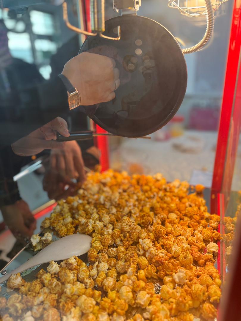 During a popcorn machine demo, two people work together—one holds the lid open while the other scoops freshly made popcorn from inside the machine.