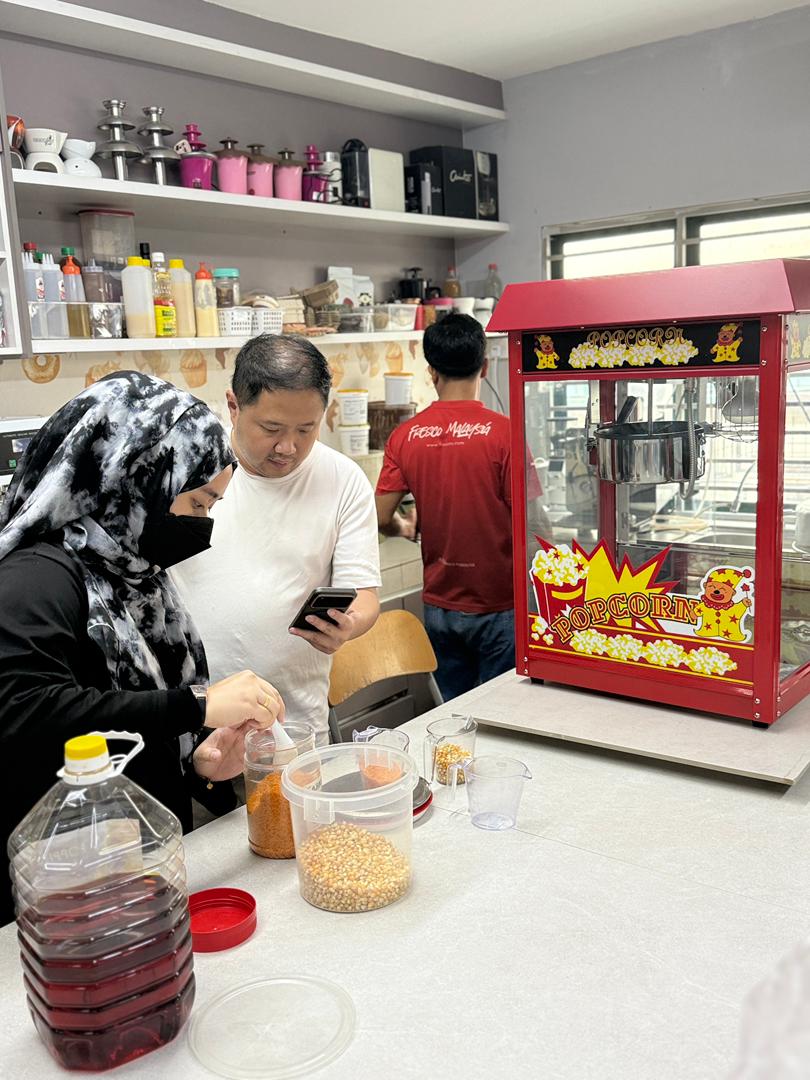 Two people prepare popcorn ingredients at a counter beside a popcorn machine, while another person in a red shirt stands nearby, participating in a machine tutorial within a kitchen setting.