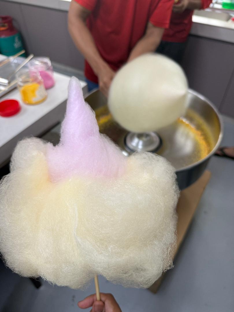 A person holds a stick of cotton candy, while in the background, someone in a red shirt demonstrates candy floss machine training and prepares another fluffy treat.