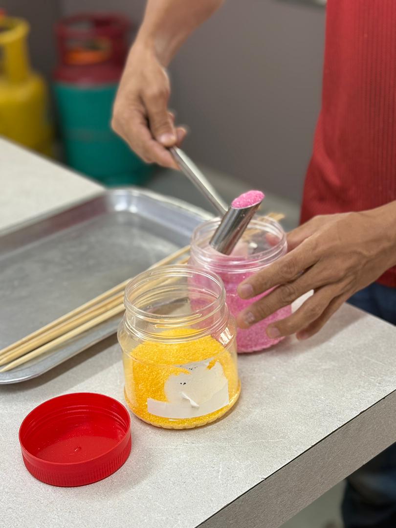 A person follows a candy floss machine training by dipping a metal mold into a jar of pink sugar crystals, with yellow sugar crystals and wooden skewers nearby on the countertop.