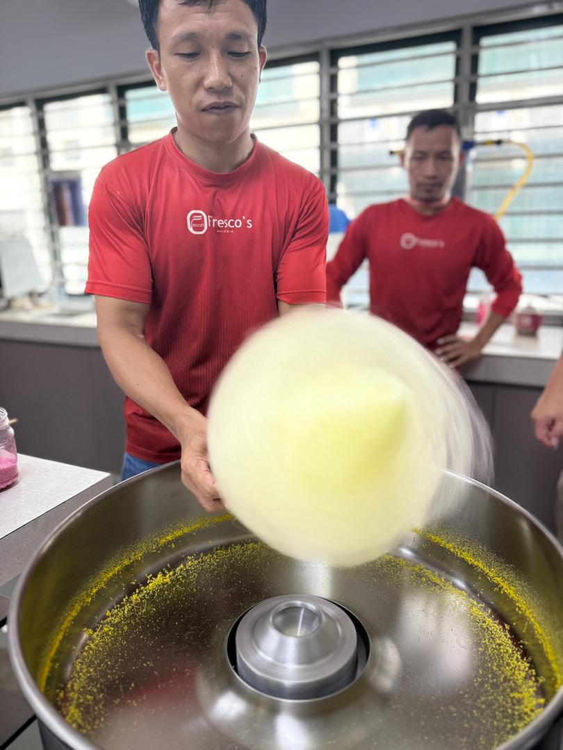 A man in a red shirt operates a cotton candy machine, spinning yellow candy floss as part of a popcorn machine training, while another man in a similar shirt stands nearby in the bright room.