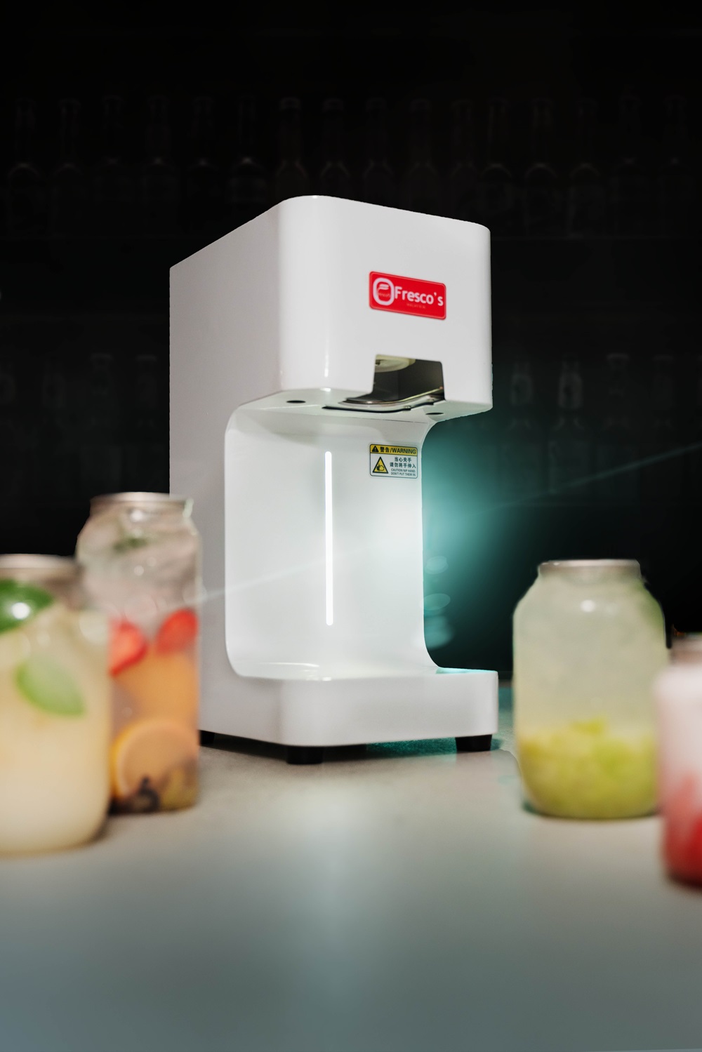 A white Fresco's beverage machine on a counter, surrounded by glass jars with various colorful drinks and a drink sealing machine, set against a dark background.