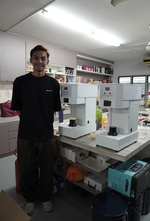 A man stands in a kitchen next to two white countertop machines, including a can bottle sealing machine, with shelves and various kitchen items in the background.