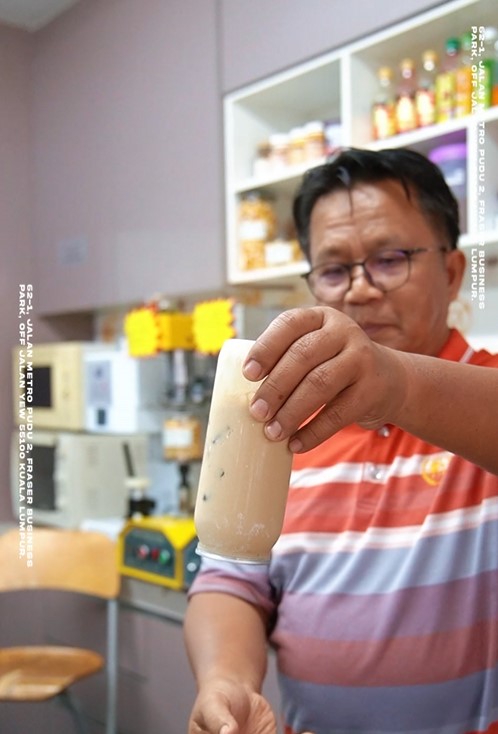 A man in a striped shirt holds up a plastic bag containing a beige-colored beverage, standing in a shop with shelves, equipment, and a drink sealer visible in the background.