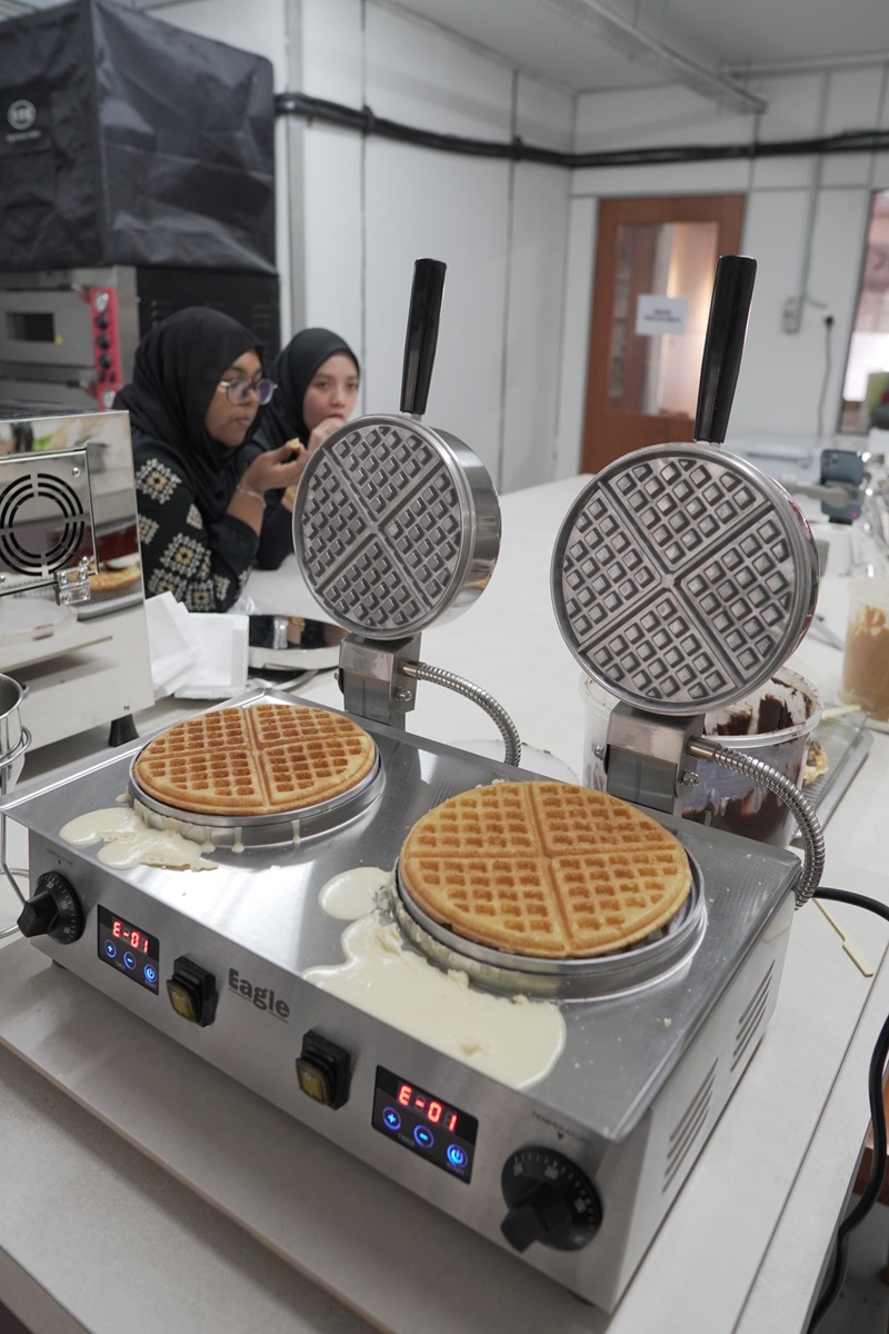 Two crispy waffles cook on the best waffle machine, batter spilling over the edges; two women in hijabs converse in the background.