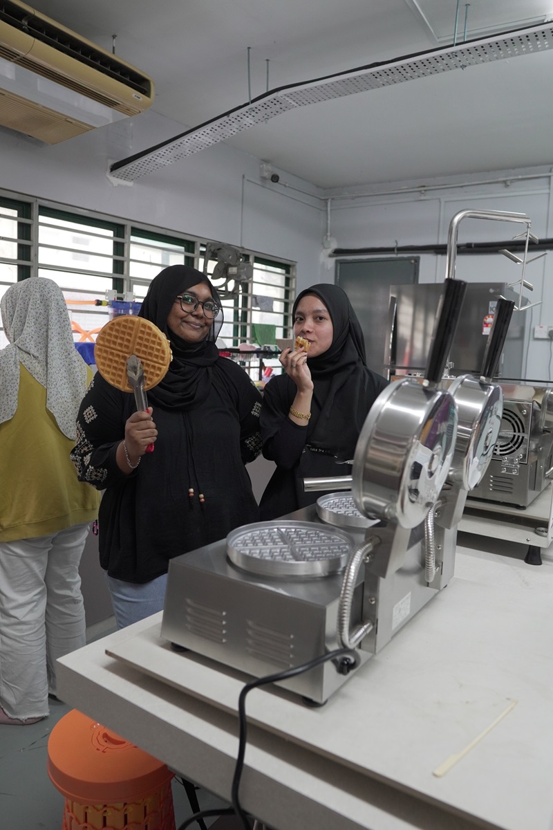 Two women in hijabs stand in a kitchen, one holding a crispy waffle and the other eating. A commercial waffle machine sits on the counter, with another person visible in the background.
