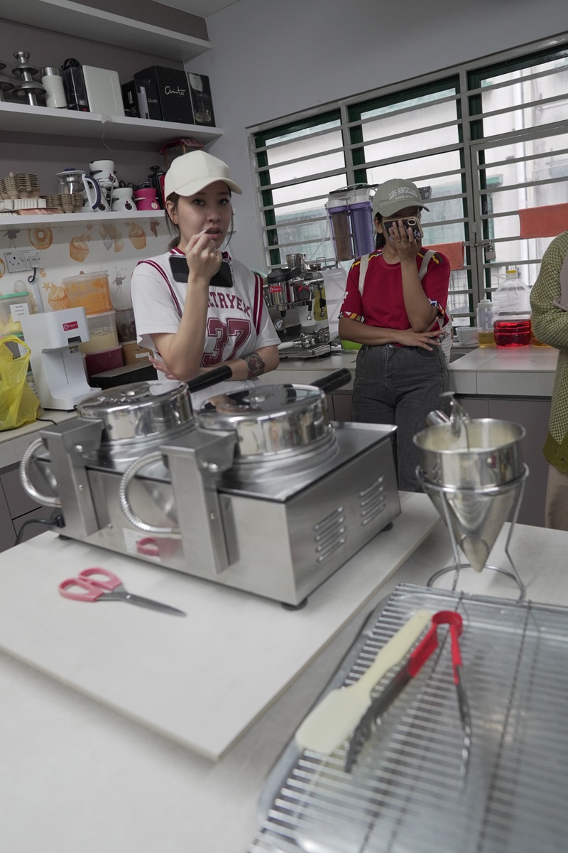 Two people in casual clothes stand in a kitchen next to cooking equipment, one enjoying a crispy waffle while the other looks at a phone. Various utensils, ingredients, and an Eagle Waffle Machine are on the counter.