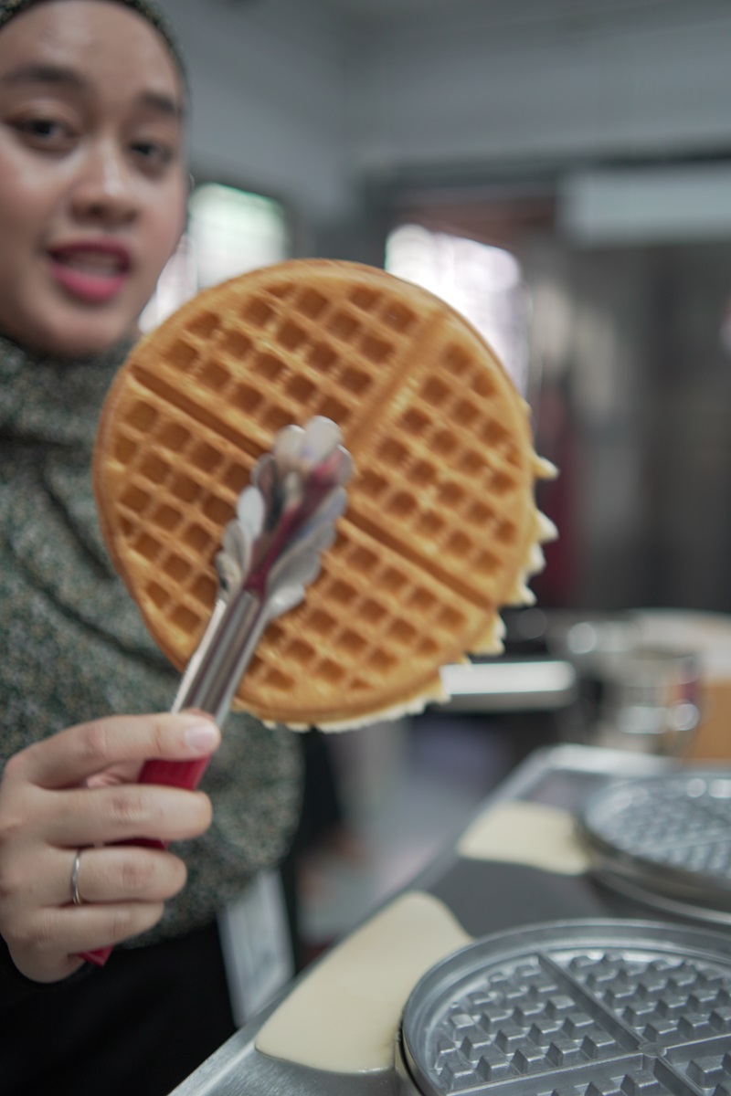 A person holds a freshly cooked round waffle with tongs in a kitchen setting, with an Eagle waffle maker and other commercial waffle machines visible in the foreground.