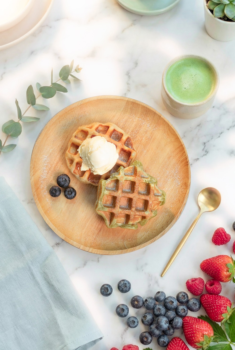 Two crispy fluffy Belgian waffles, one topped with a scoop of ice cream, are served on a wooden plate with blueberries. Fresh berries, a cup of green tea, and a gold spoon are arranged nearby on the marble surface.